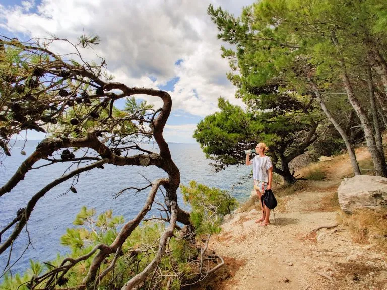 Joven turista activo tomando un descanso, bebiendo agua, llevando una pequeña mochila mientras camina por el sendero costero entre pinos en busca de una cala remota para nadar solo en paz junto al mar en Croacia.